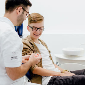 Ein Kieferorthopäde erklärt einem Jugendlichen mit Zahnspange die Zahnpflege im Behandlungsraum.