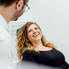 Frau sitzt lachend im Zahnarztstuhl bei Beratung in moderner Zahnarztpraxis.