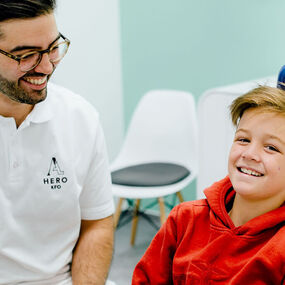 Lächelnder Zahnarzt mit Brille begrüßt fröhlichen Jungen auf Behandlungsstuhl in moderner Praxis.