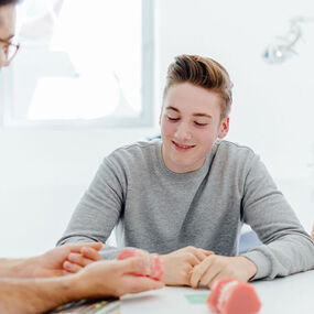 Kieferorthopäde erklärt Jugendlichen und Mutter mit Zahnmodell die Zahnspangenbehandlung.